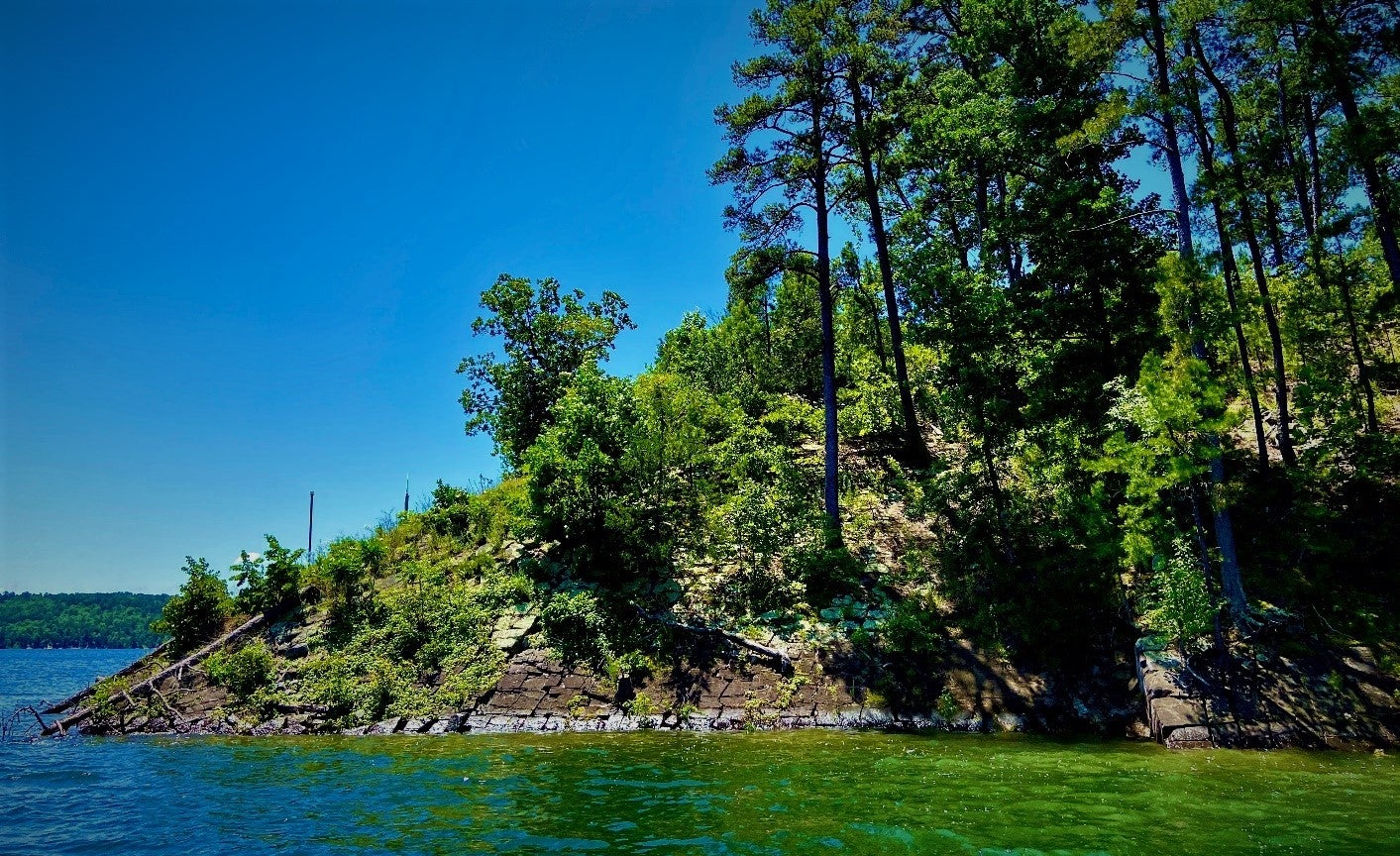 An island covered in green trees sits in a lake. Dark gray rocks line the shore and are arranged in a checkboard pattern. 
